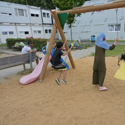 Zwei Kinder sitzen auf einer Schaukel. Eine Mutter steht dabei. Im Hintergrund sieht man die grauen Containerbauten.