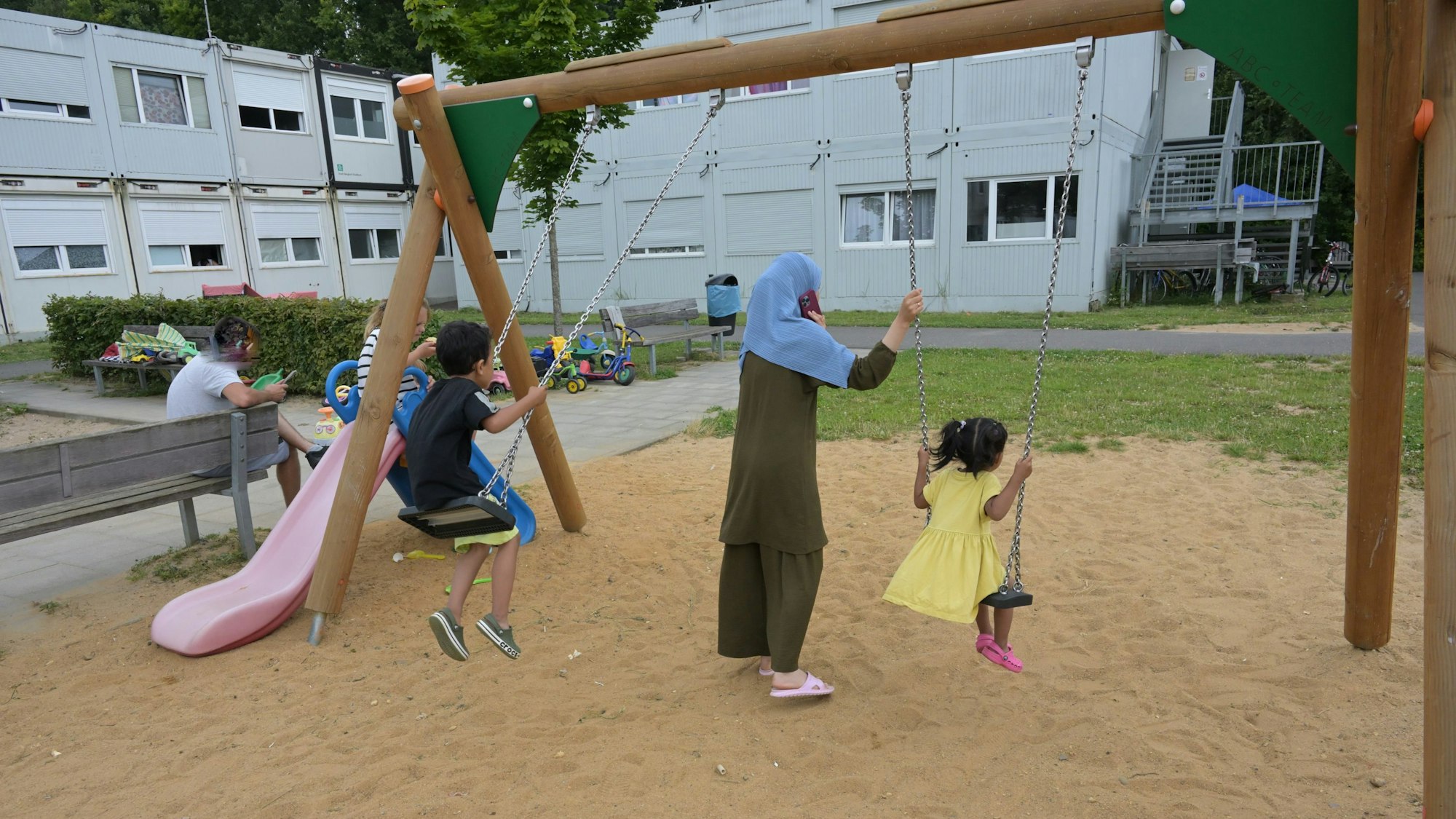 Zwei Kinder sitzen auf einer Schaukel. Eine Mutter steht dabei. Im Hintergrund sieht man die grauen Containerbauten.
