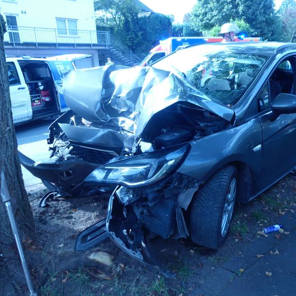 Ein Unfallauto, das nach einem Aufprall gegen einen Baum an der Frontseite völlig zerstört ist. Im Hintergrund sind Rettungskräfte zu sehen.