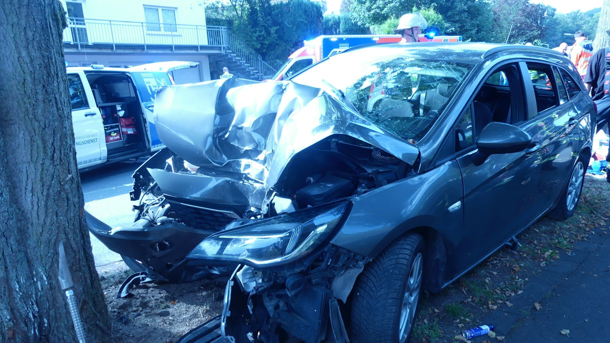 Ein Unfallauto, das nach einem Aufprall gegen einen Baum an der Frontseite völlig zerstört ist. Im Hintergrund sind Rettungskräfte zu sehen.