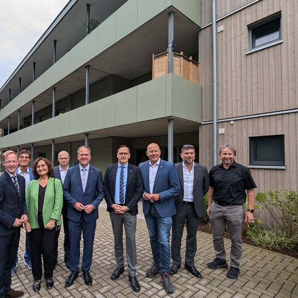 Das Foto zeigt eine Gruppe vor dem Mehrfamilienhaus mit Holzfassade.