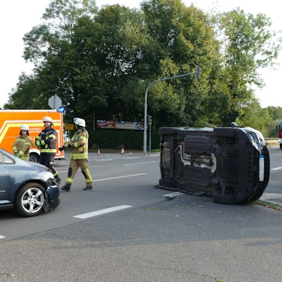 Ein schwarzes Auto liegt auf der Seite auf einer Kreuzung in Lohmar, Feuerwehrleute stehen auf der Straße.