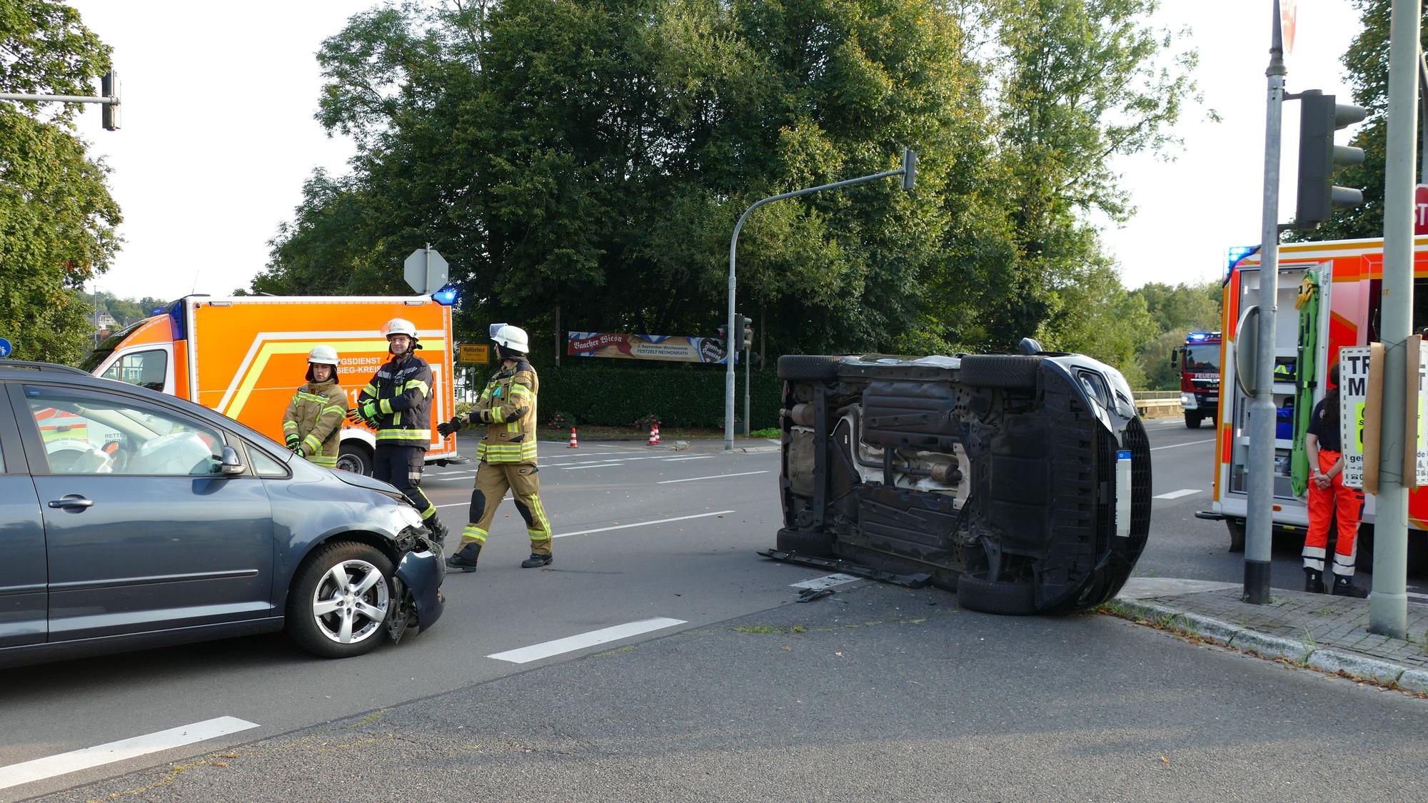 Ein schwarzes Auto liegt auf der Seite auf einer Kreuzung in Lohmar, Feuerwehrleute stehen auf der Straße.