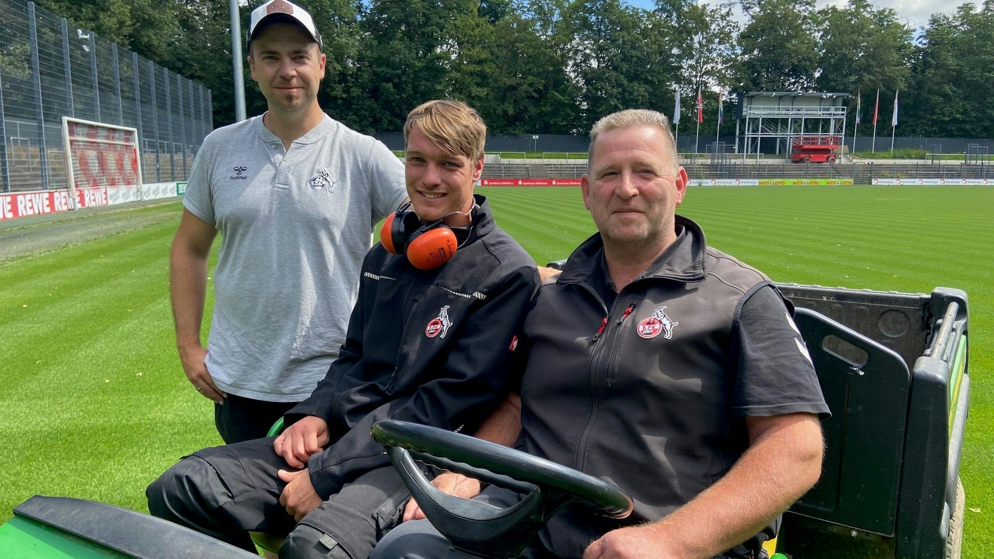 Auf dem Bild sind drei Greenkeeper auf einem Aufsitztraktor zu sehen.