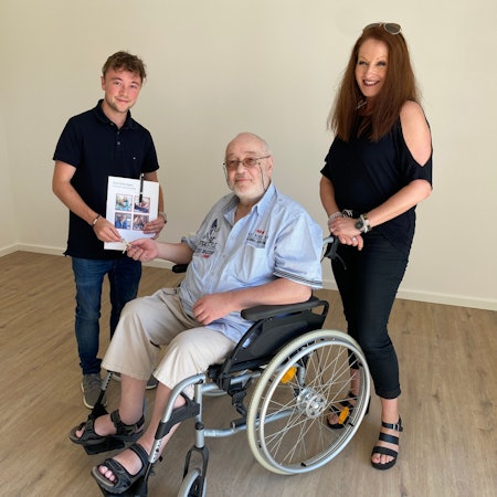 Philip Wiskirchen übergibt einen Schlüssel an Heinz Kesten, der im Rollstuhl sitzt. Kümmerin Anuschka Schwinning-Hess steht daneben.
