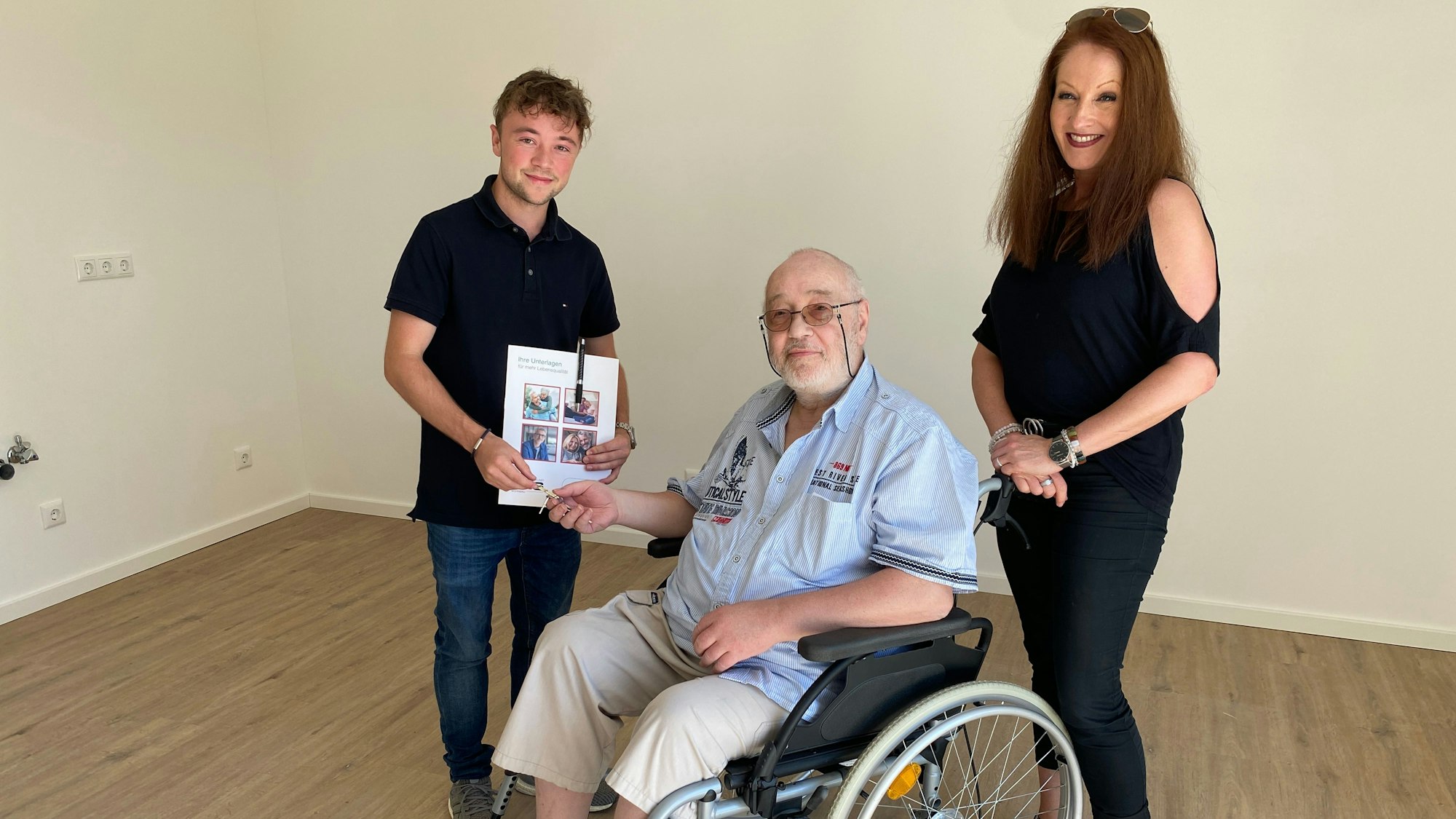 Philip Wiskirchen übergibt einen Schlüssel an Heinz Kesten, der im Rollstuhl sitzt. Kümmerin Anuschka Schwinning-Hess steht daneben.