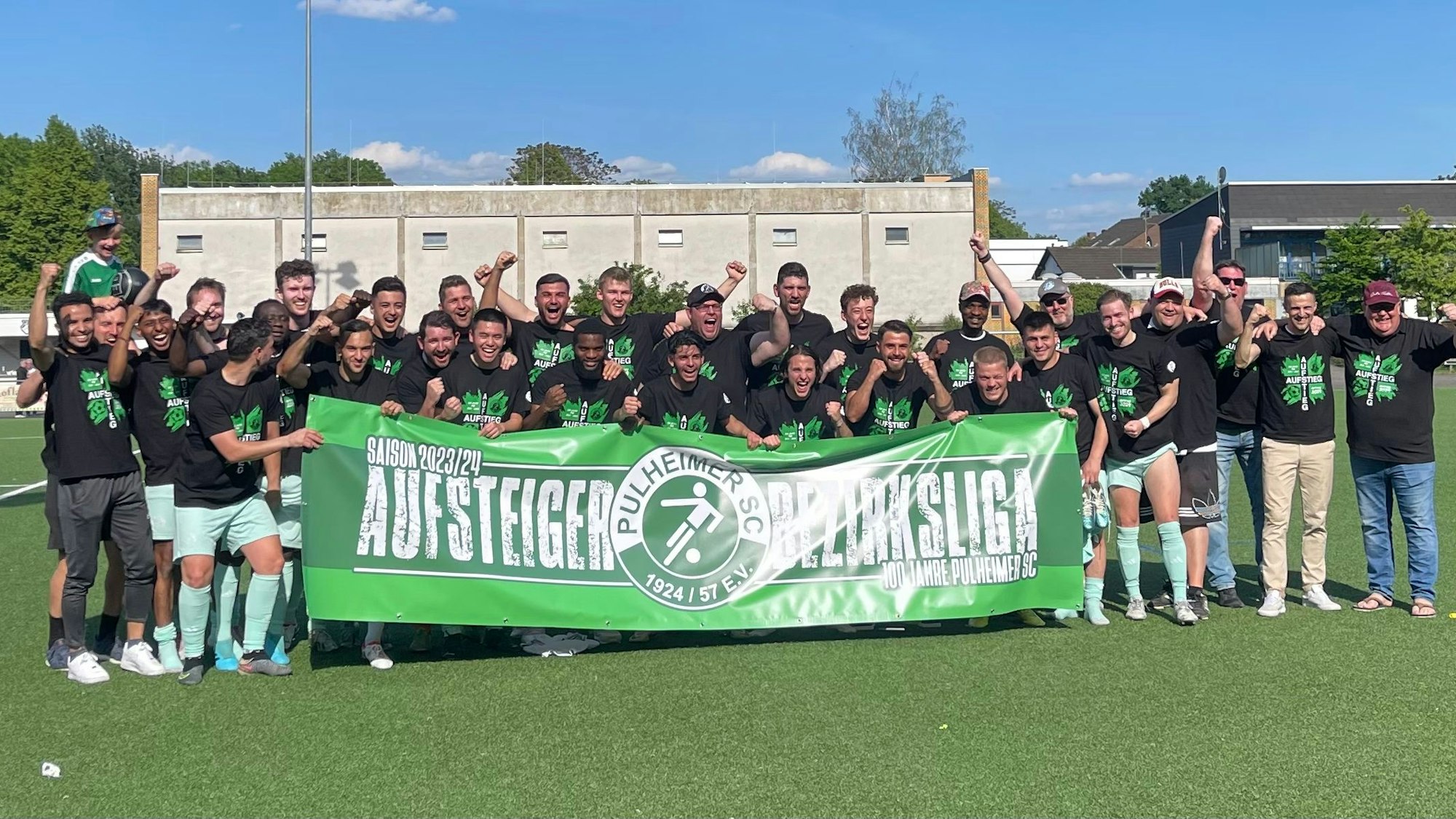 Die Spieler des Pulheimer SC tragen schwarze TShirts mit einem grünen Logo. Sie halten ein Banner mit der Aufschrift: Saison 2023/24. Aufstieg Bezirksliga. 100 Jahre Pulheimer SC.
