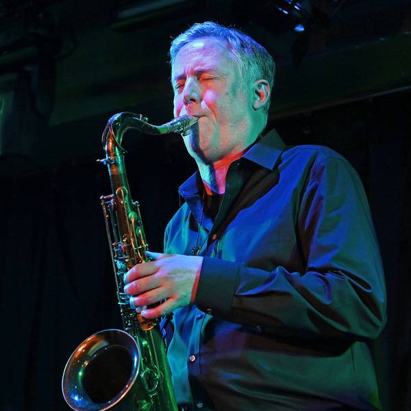 Der Musiker trägt ein dunkles Hemd und bläst in sein Saxofon, der Hintergrund ist dunkel. Vor ihm ein längliches Mikrofon.
