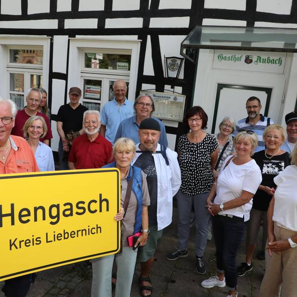 Michael Iwanzik aus Kürten-Olpe hält ein Ortsschild mit der Aufschrift „Hengasch Kreis Liebernich“ und steht mit einer Gruppe vor dem Gasthof Aubach der Schmunzel-Krimiserie „Mord mit Aussicht“.