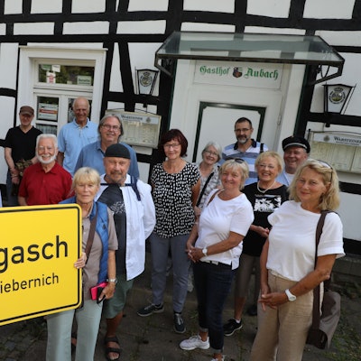 Michael Iwanzik aus Kürten-Olpe hält ein Ortsschild mit der Aufschrift „Hengasch Kreis Liebernich“ und steht mit einer Gruppe vor dem Gasthof Aubach der Schmunzel-Krimiserie „Mord mit Aussicht“.
