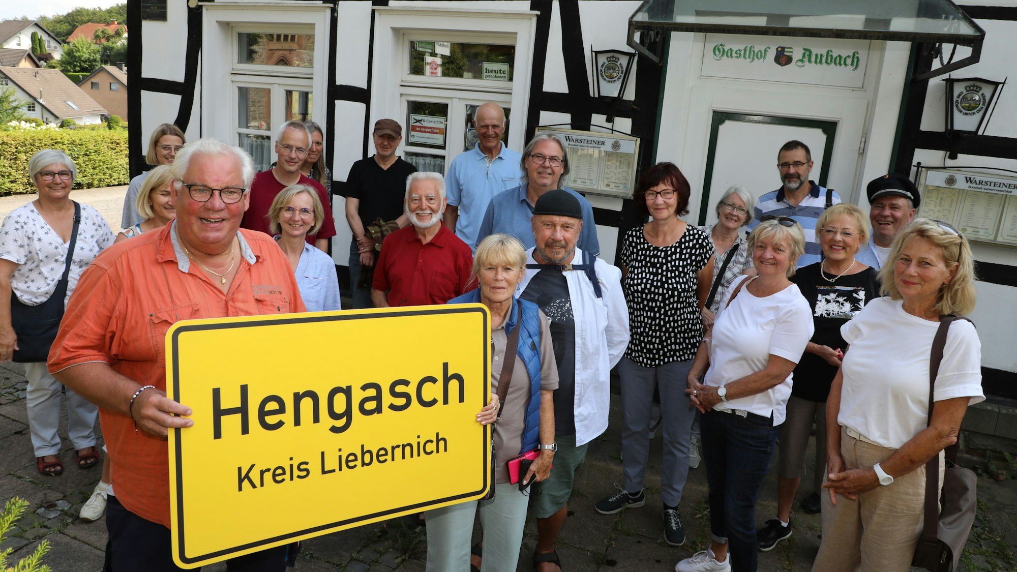 Michael Iwanzik aus Kürten-Olpe hält ein Ortsschild mit der Aufschrift „Hengasch Kreis Liebernich“ und steht mit einer Gruppe vor dem Gasthof Aubach der Schmunzel-Krimiserie „Mord mit Aussicht“.