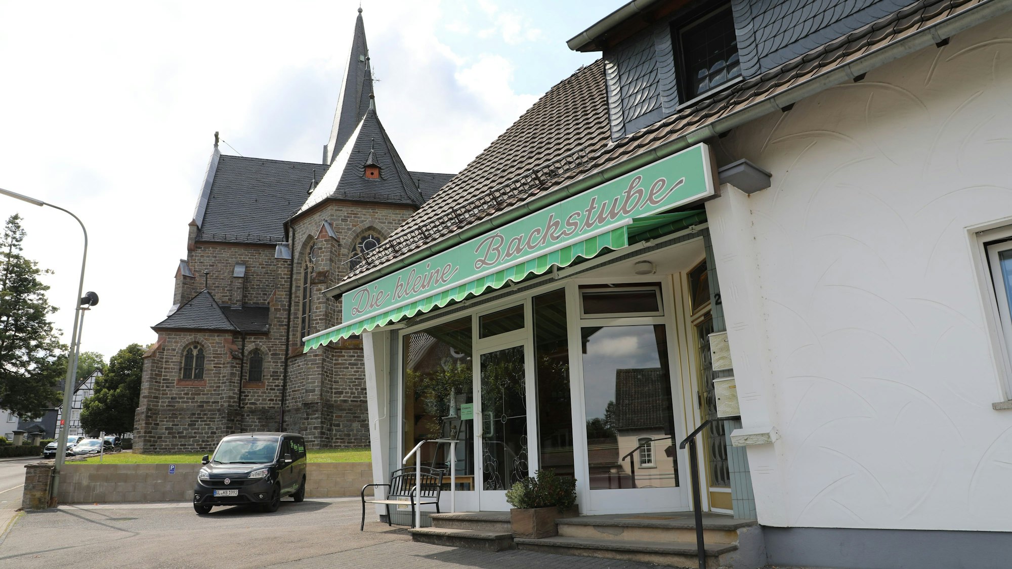 Vor einer Kirche ist ein Laden mit der Aufschrift „Die kleine Backstube“ im Ortskern von Kürten-Olpe zu sehen.