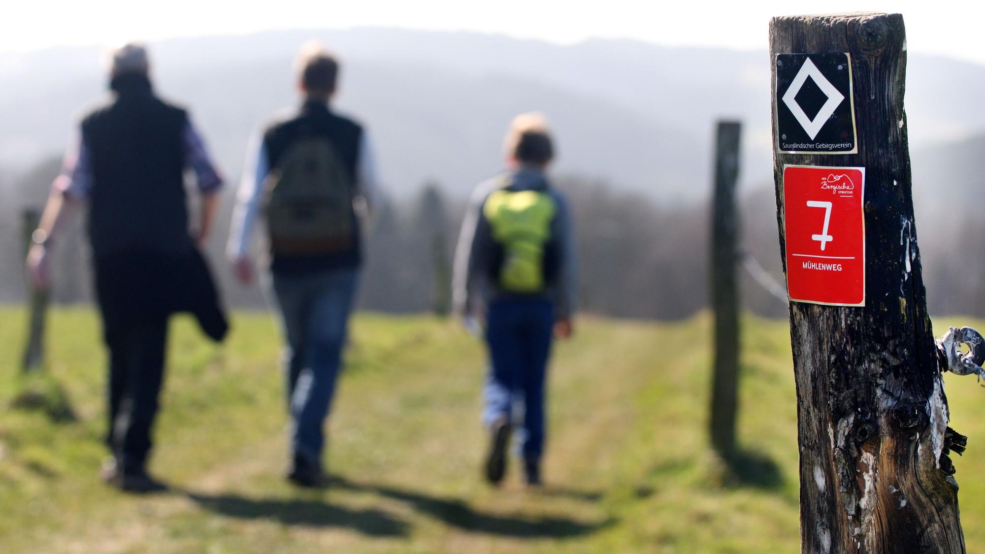 Eine Familie wandert auf dem Kürtener Mühlenweg, an einem Zaunpfosten ist eine Wegemarkierung zu sehen.