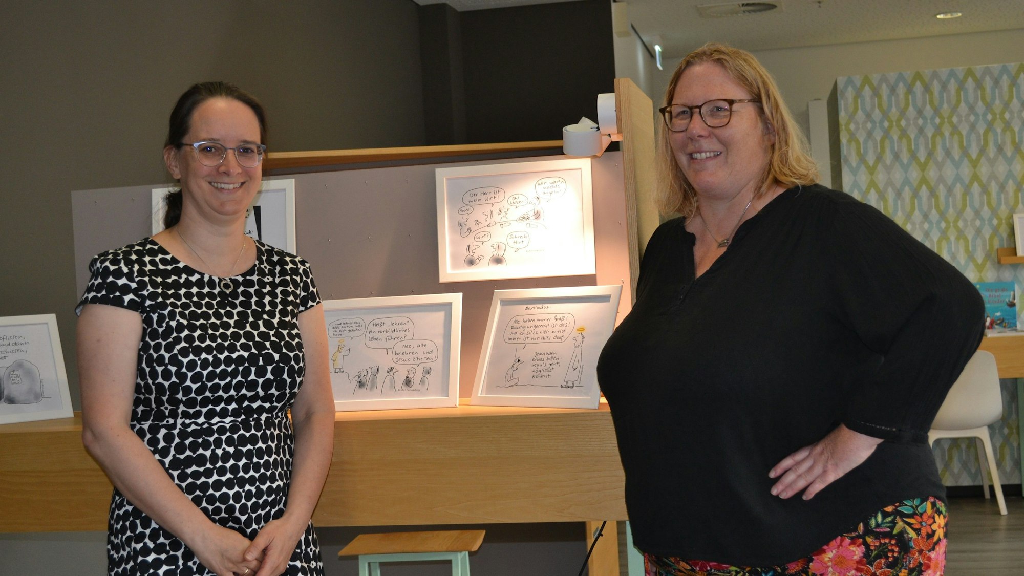 Zwei Frauen stehen vor einem Tisch mit gerahmten Zeichnungen.