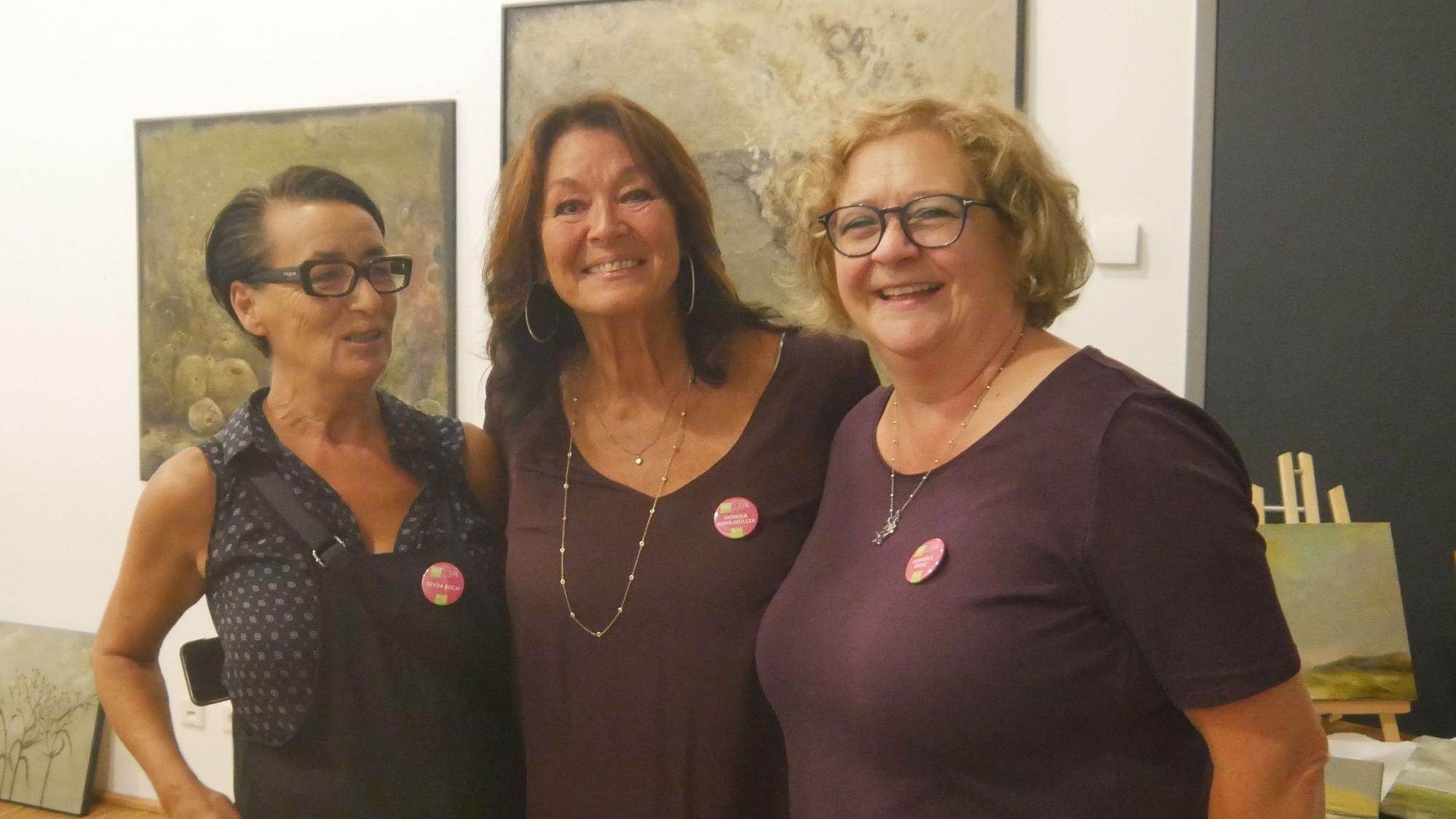 Drei Frauen stehen eng beieinander in einer Kunstausstellung.