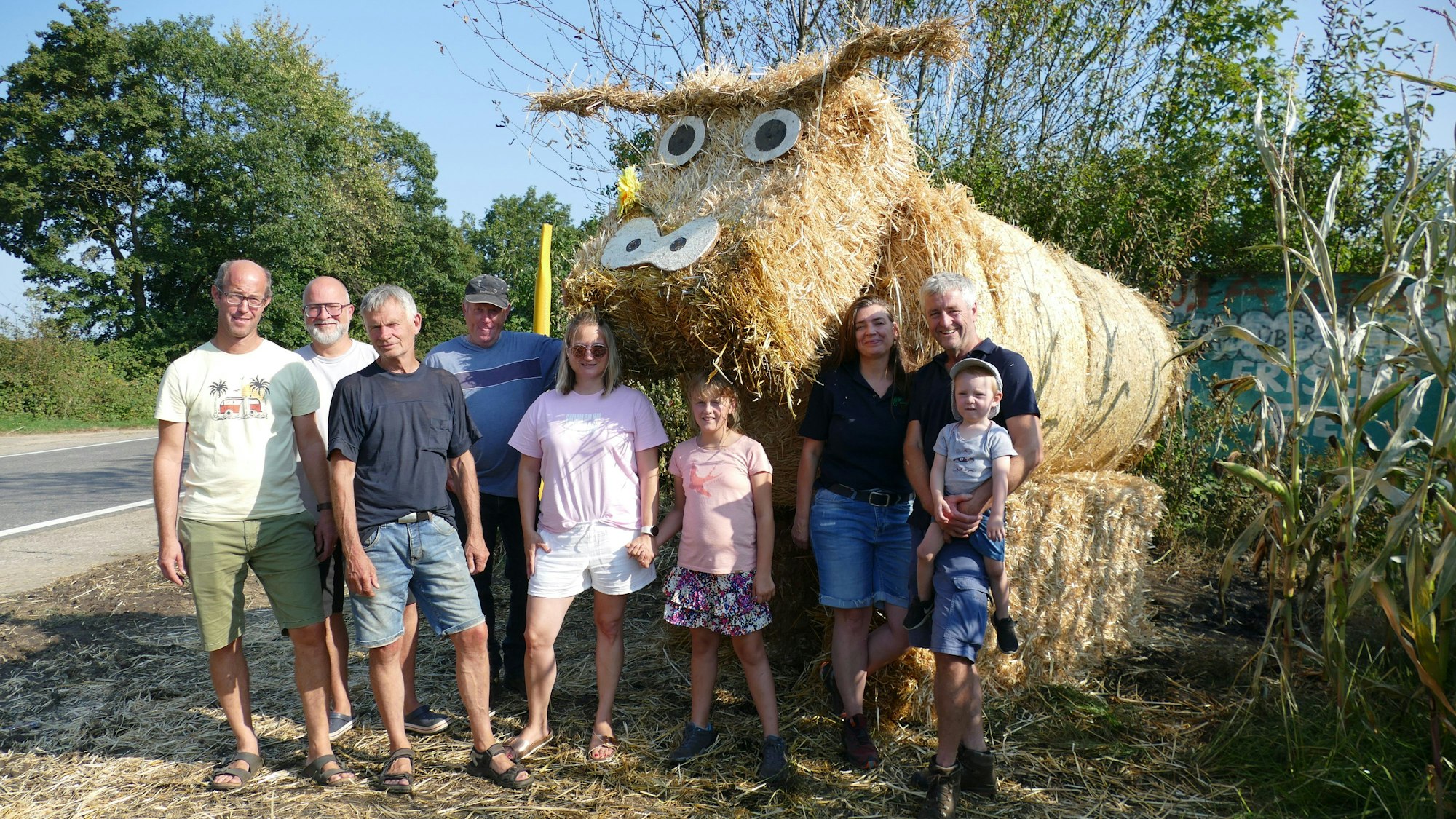 Männer, Frauen und Kinder stehen vor einer Kuh aus Strohballen.