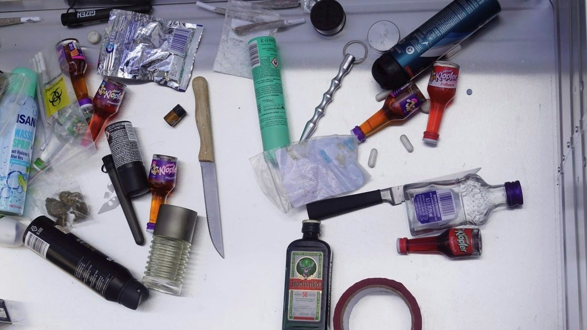 Das Bild zeigt unter anderem zwei Messer, kleinere Flaschen Alkohol, Joints und Deos.
