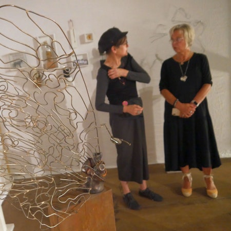 Zwei Frauen in einer Kunstausstellung. Im Vordergrund steht ein Schmetterling, der aus Draht geformt ist, auf einem hölzernen Sockel.