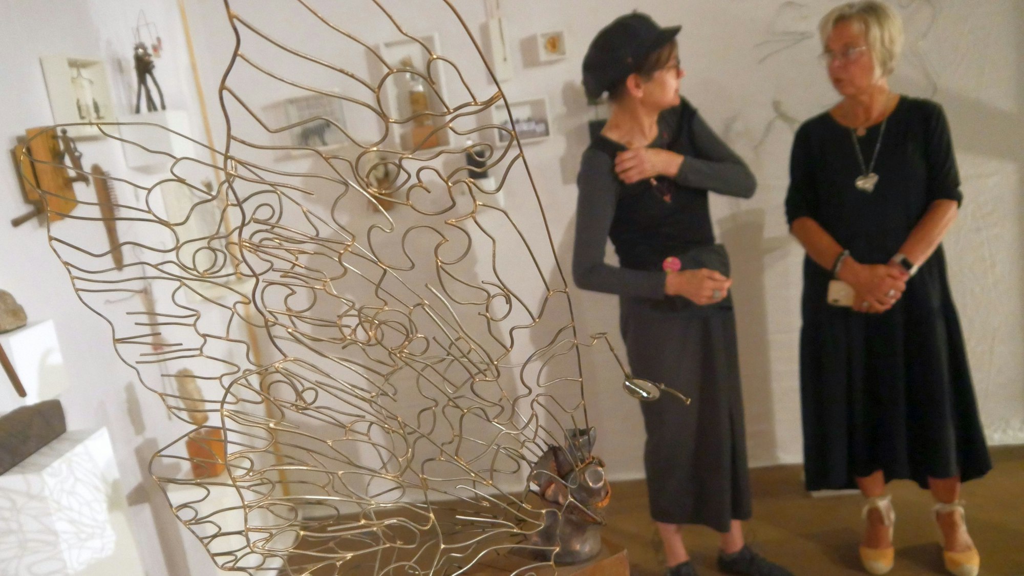 Zwei Frauen in einer Kunstausstellung. Im Vordergrund steht ein Schmetterling, der aus Draht geformt ist, auf einem hölzernen Sockel.