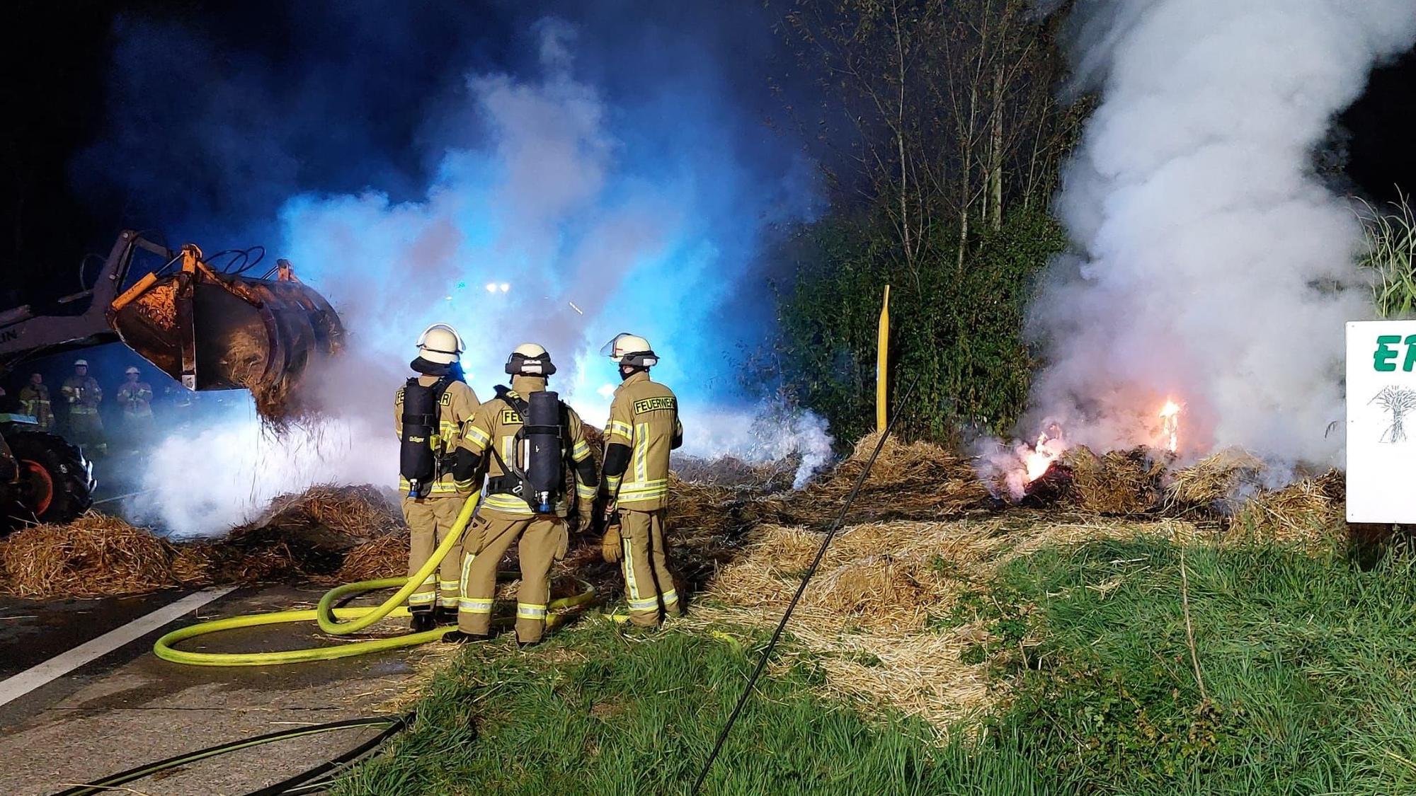 Helfer der Feuerwehr löschen Strohballen auf einer Wiese.