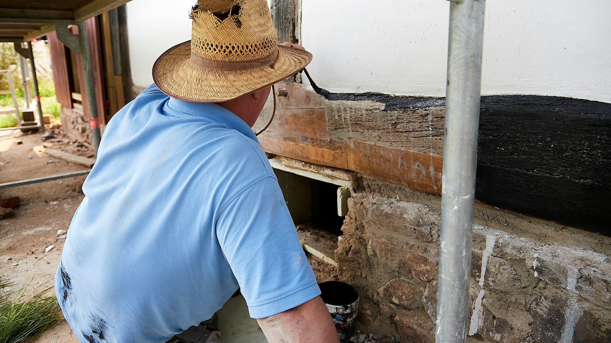 Das Bild zeigt einen Mann, der mit einem Pinsel schwarze Farbe auf einen Balken aufträgt.