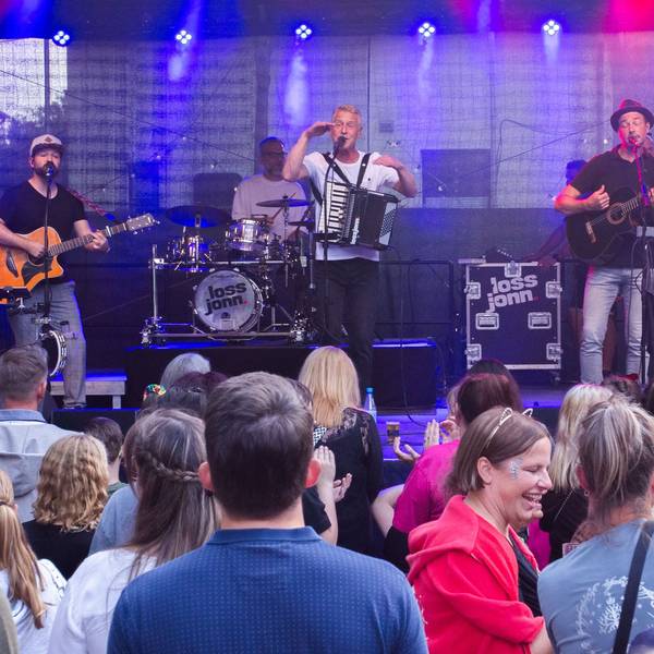 Drei Musiker mit Instrumenten stehen auf einer Bühne, viele Menschen sind davor zu sehen.