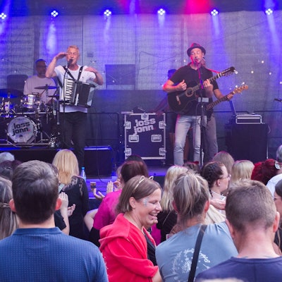 Drei Musiker mit Instrumenten stehen auf einer Bühne, viele Menschen sind davor zu sehen.