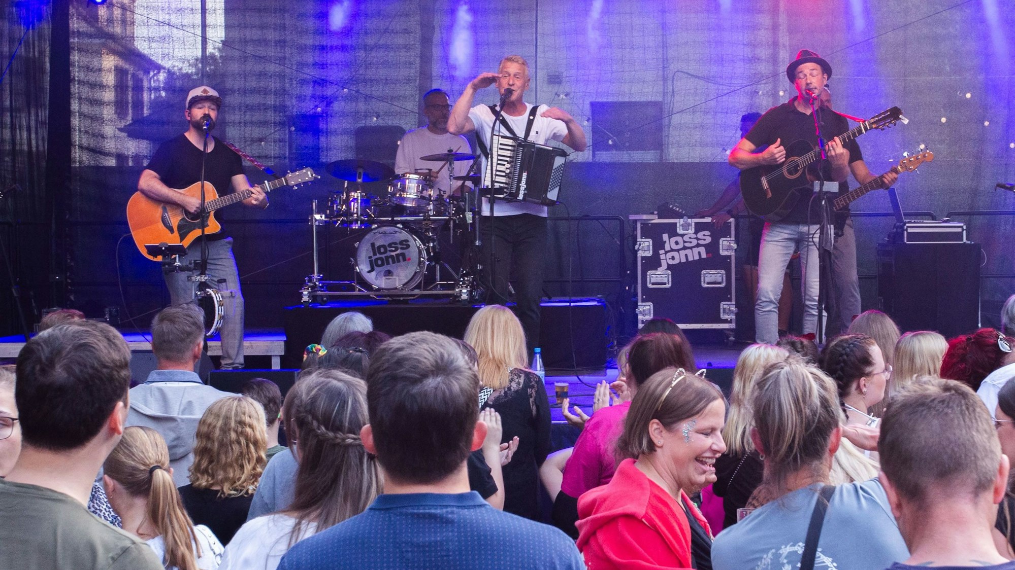 Drei Musiker mit Instrumenten stehen auf einer Bühne, viele Menschen sind davor zu sehen.