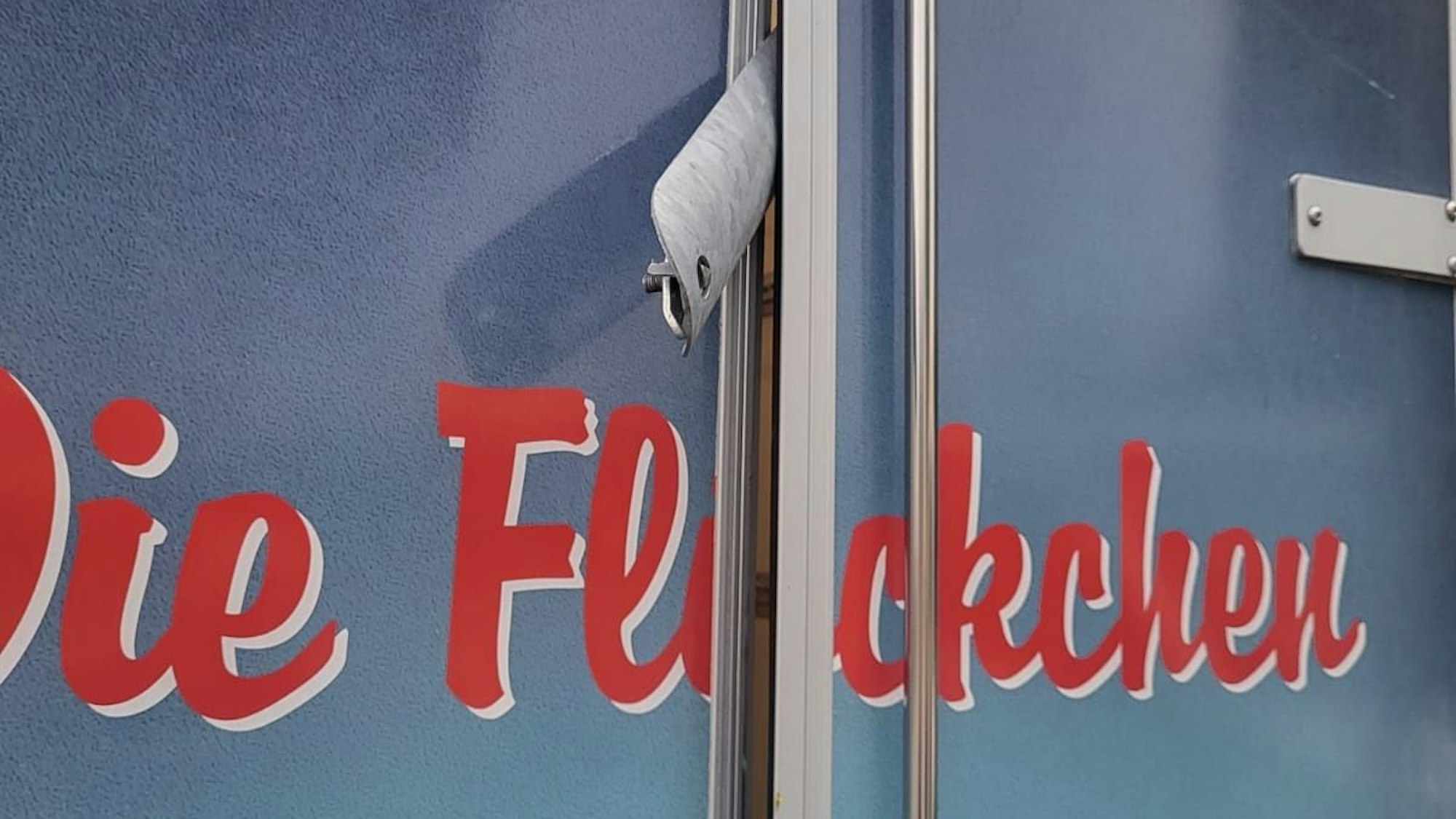 Im Türspalt eines Pkw-Anhängers mi der Aufschrift „Die Flöckchen“ steckt die Metallabdeckung einer Laternenstromzuführung.