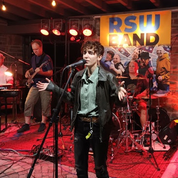 Eine Person singt am Mikrofon, umringt von Musikern