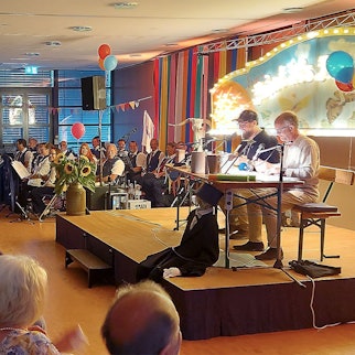 Ralf Kramp (r.) und Carsten S. Henn sitzen auf der Bühne, davor das Publikum. Die Musikkapelle Nettersheim ist hinten links platziert.