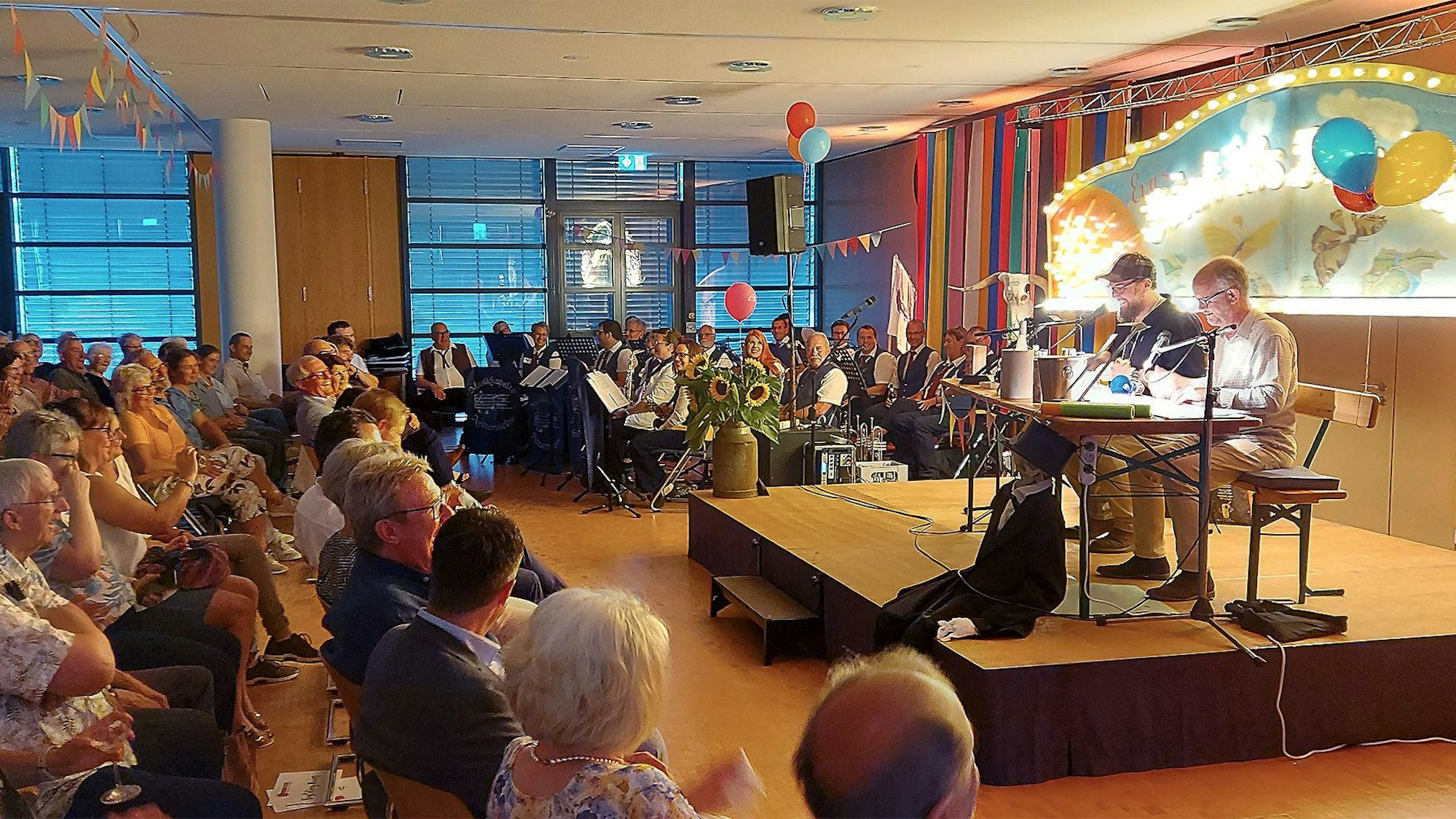 Ralf Kramp (r.) und Carsten S. Henn sitzen auf der Bühne, davor das Publikum. Die Musikkapelle Nettersheim ist hinten links platziert.