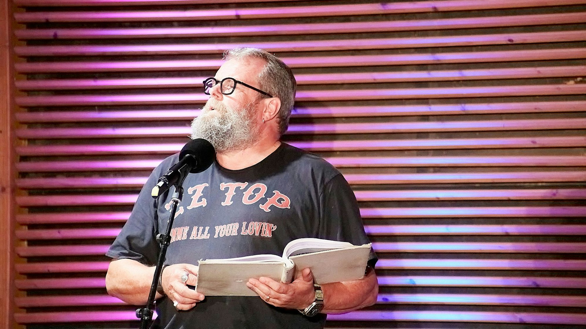 Jochen Malmsheimer steht im T-Shirt der Band ZZ Top am Mikrofon auf einer Bühne und hält ein aufgeschlagenes Buch in den Händen.