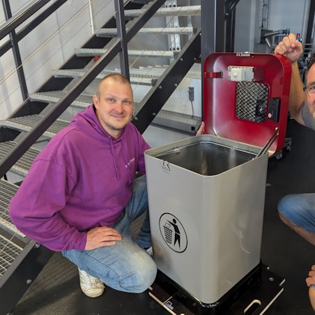 Das Foto zeigt zwei Männer, die neben einem Mülleimer hocken. Im geöffneten Deckel sind Sensoren zu erkennen.