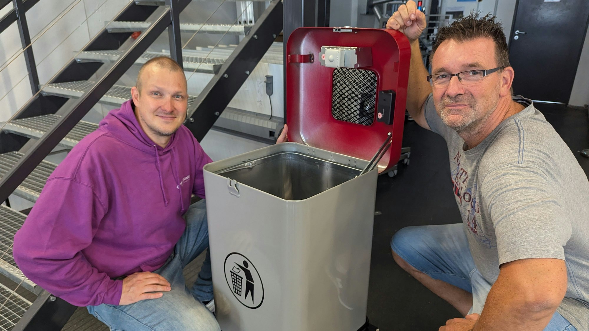 Das Foto zeigt zwei Männer, die neben einem aufgeklappten Mülleimer mit Sensor im Deckel hocken.