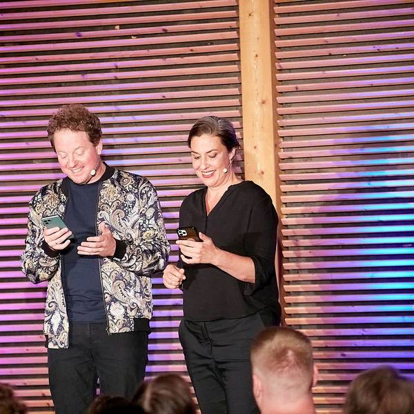 Eva Eiselt und Martin Zingsheim schauen grinsend auf ihre Handys, während sie auf einer Bühne vor Publikum stehen.