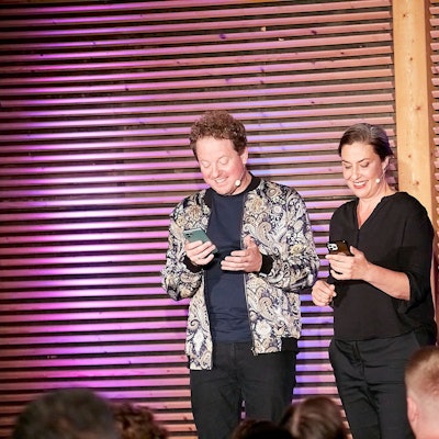 Eva Eiselt und Martin Zingsheim schauen grinsend auf ihre Handys, während sie auf einer Bühne vor Publikum stehen.