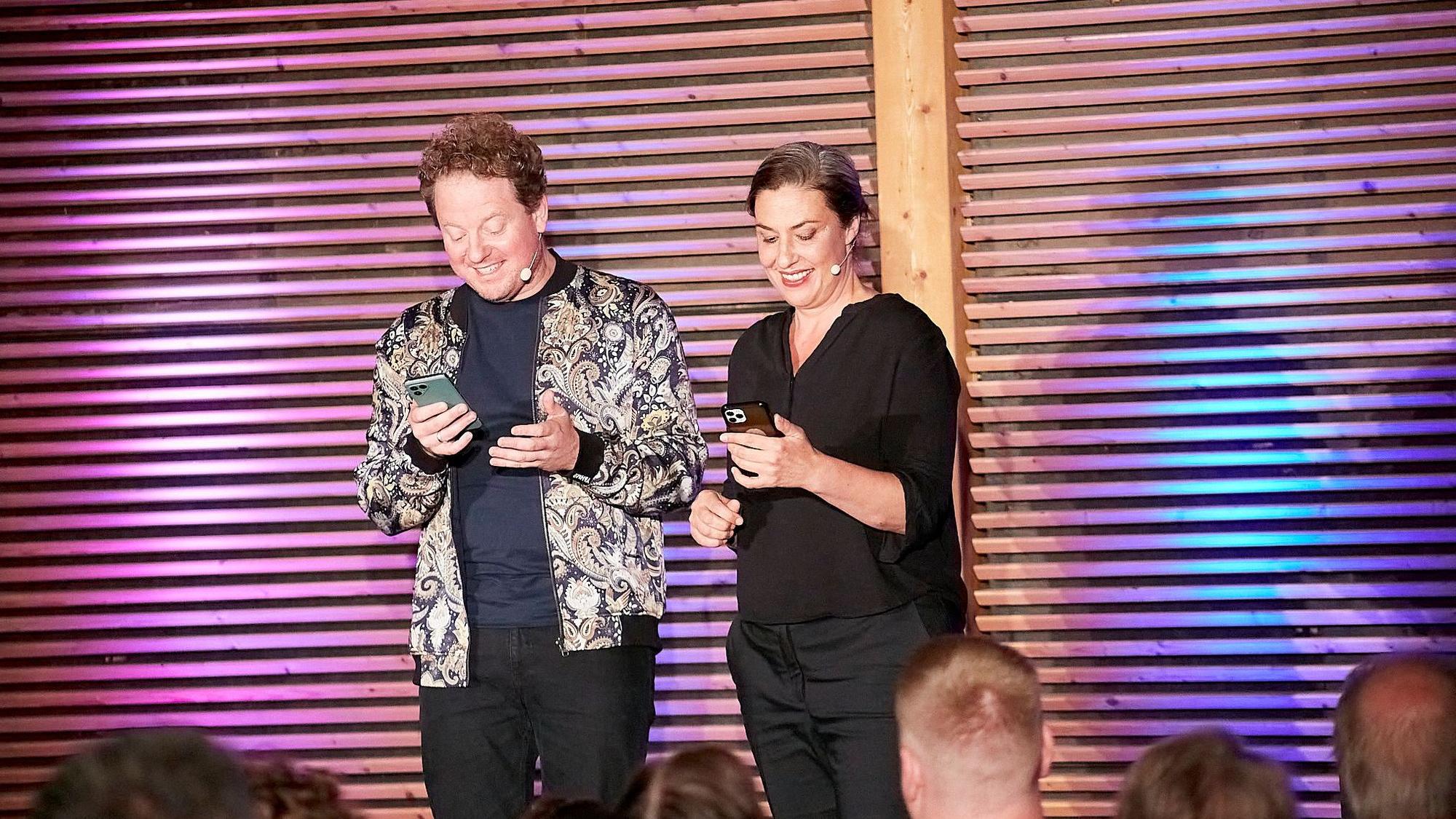 Eva Eiselt und Martin Zingsheim schauen grinsend auf ihre Handys, während sie auf einer Bühne vor Publikum stehen.