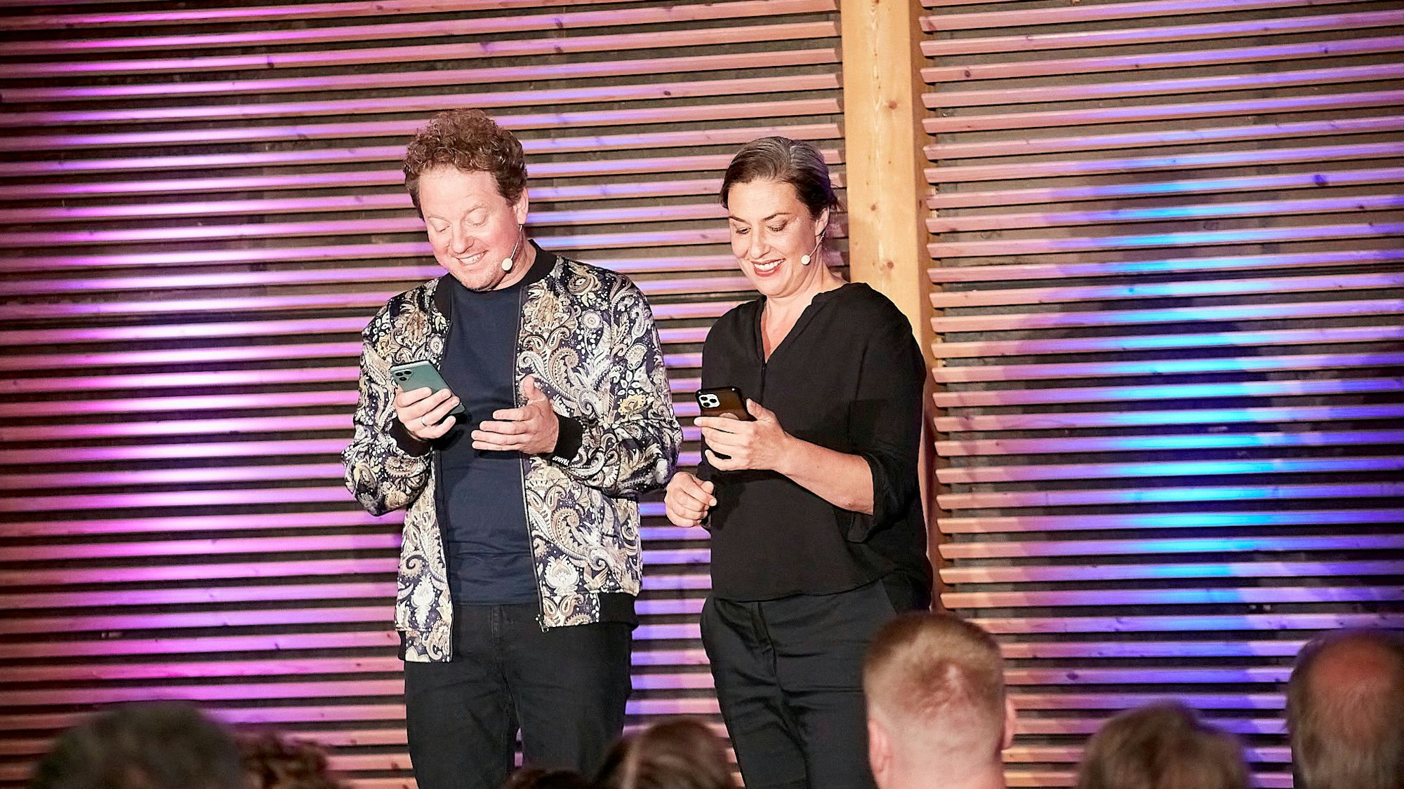 Eva Eiselt und Martin Zingsheim schauen grinsend auf ihre Handys, während sie auf einer Bühne vor Publikum stehen.