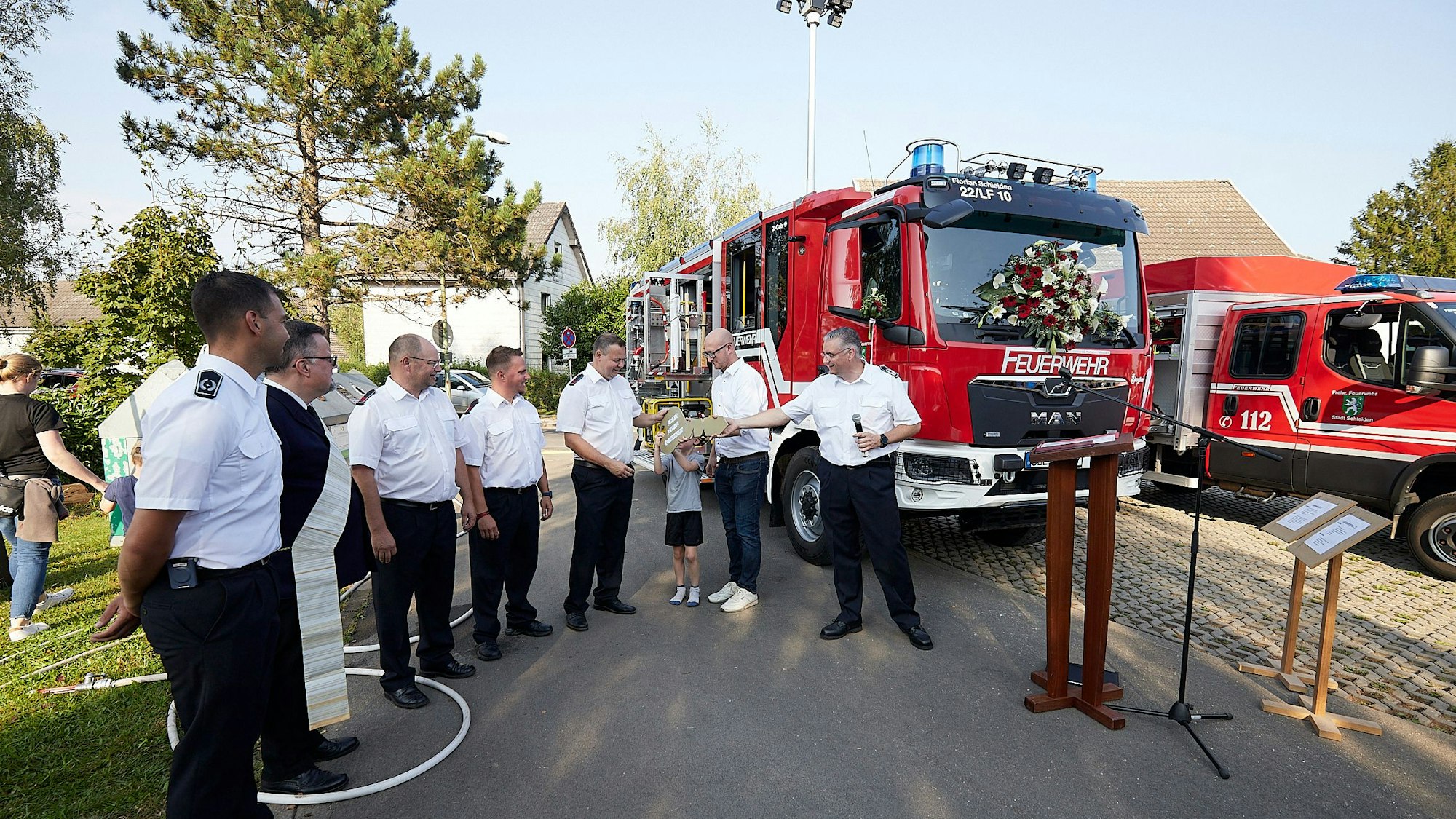 Mehrere Feuerwehrleute, ein Geistlicher, ein Kind und der Bürgermeister stehen vor einem der Feuerwehrfahrzeuge in Schleiden-Herhahn.