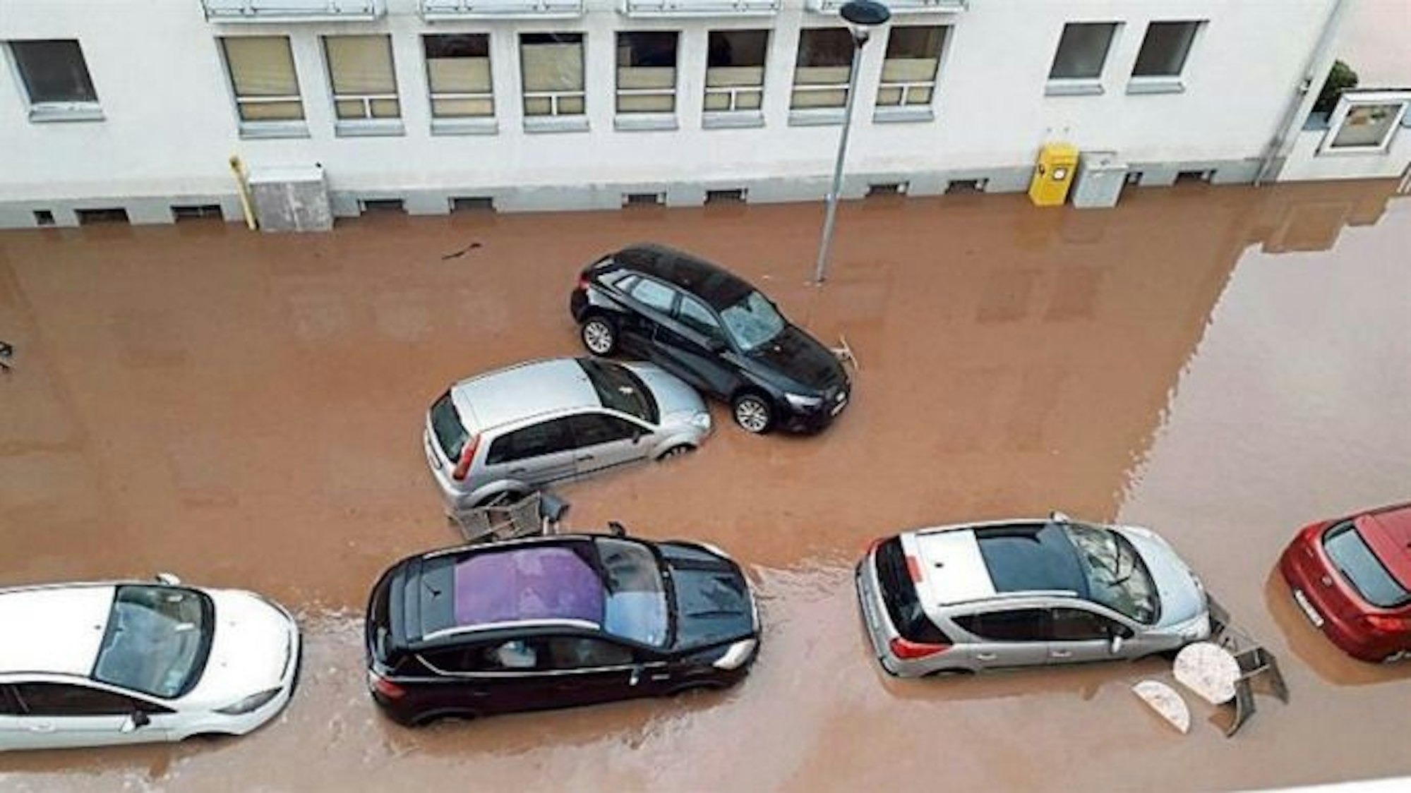 Die Straße ist überflutet. Autos stehen tief im braunen Wasser. Ein Wagen ist auf einen anderen geschoben worden.