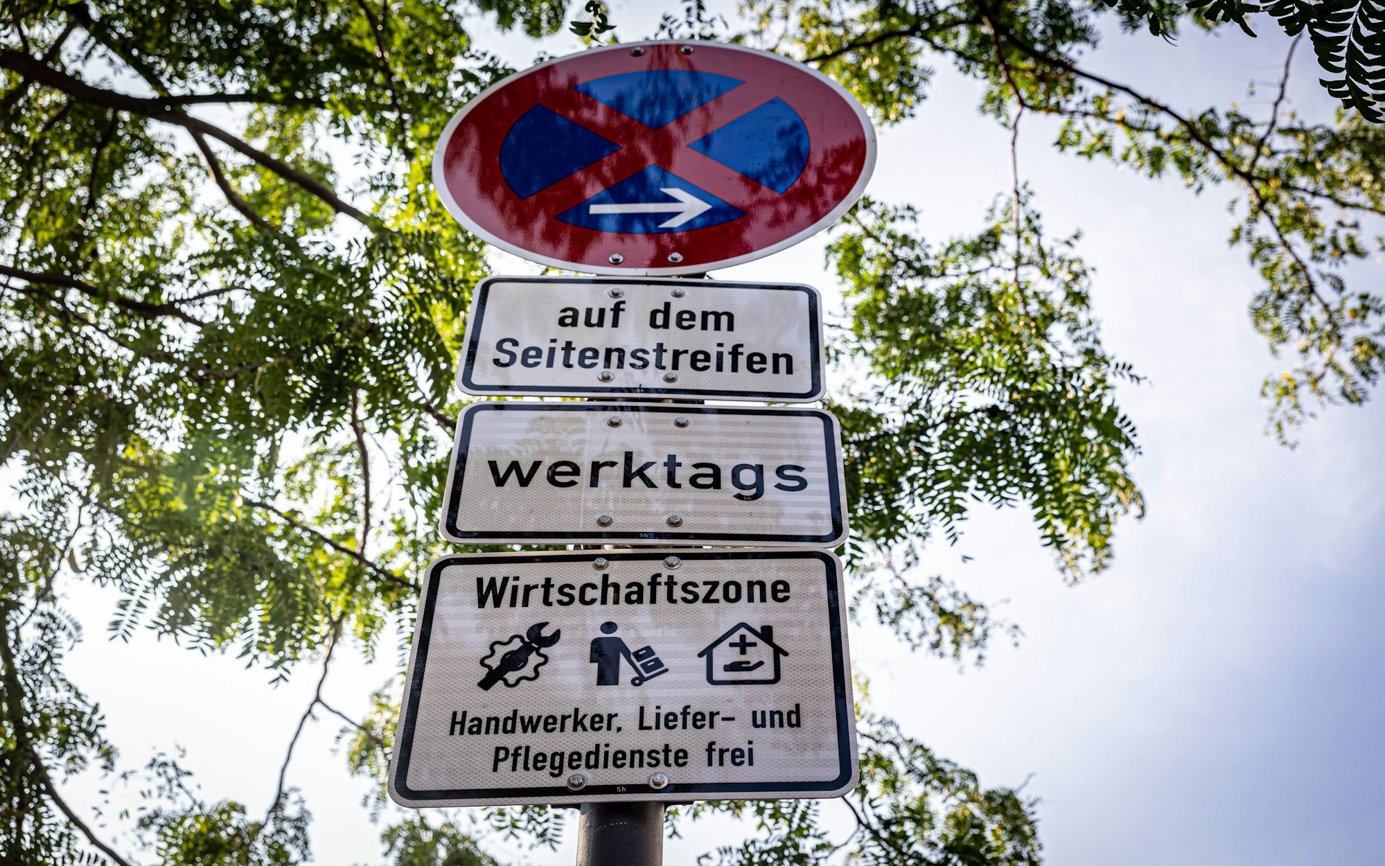 Mehrere Verkehrsschilder hängen übereinander, ein Parkverbotschild, eins, auf dem „auf dem Seitenstreifen“ steht, ein weiteres mit dem Wort werktags und eins, das auf die Wirtschaftszone hinweist.