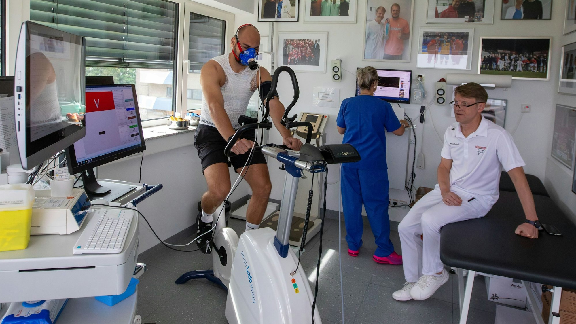 KEC-Spieler Maximilian Glötzl (l.) auf dem Cardio-Rad. Eduard Gorr, Leiter der Sportkardiologie, schaut sich die Werte auf dem Monitor an. Schwester Daniela Knappmeier im Hintergrund.