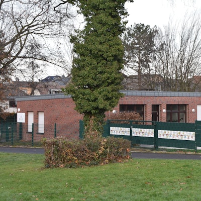 Auf dem Bild ist das Gebäude der Kita zu sehen, es besteht aus roten Backsteinen und hat ein flaches Dach. Davor ein grüner Zaun.