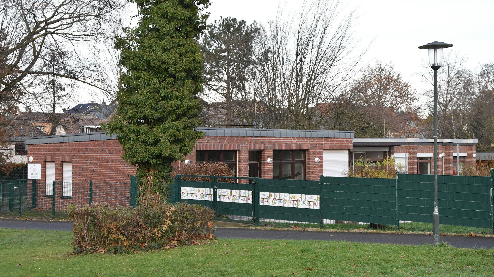Auf dem Bild ist das Gebäude der Kita zu sehen, es besteht aus roten Backsteinen und hat ein flaches Dach. Davor ein grüner Zaun.