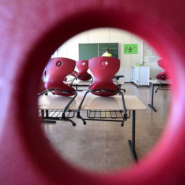 Stühle sind in einem Klassenzimmer auf Tische aufgereiht, im Hintergrund bereitet eine Lehrerin einen Tafelanschrieb vor.