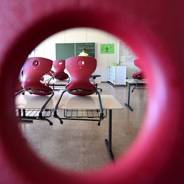 Stühle sind in einem Klassenzimmer auf Tische aufgereiht, im Hintergrund bereitet eine Lehrerin einen Tafelanschrieb vor.