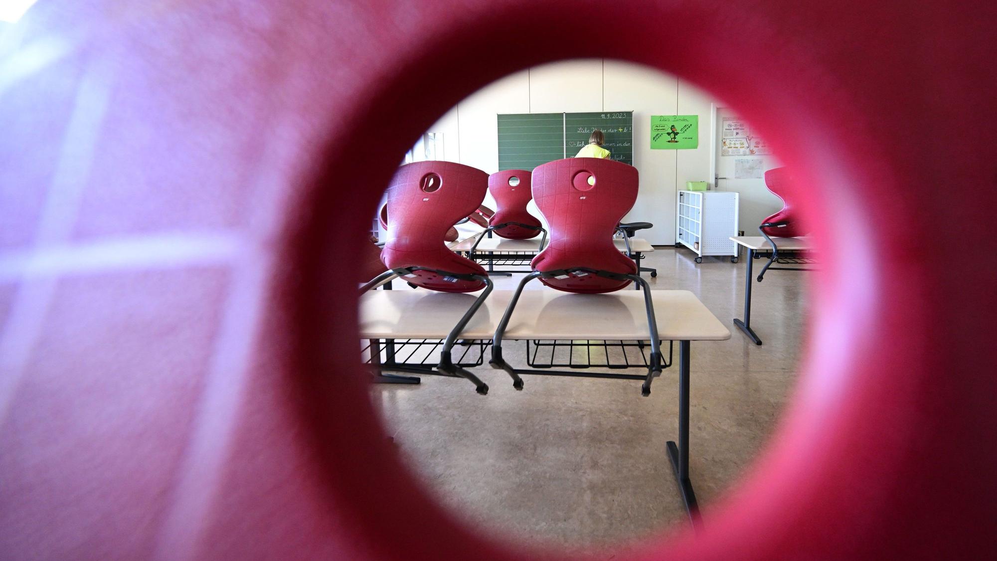 Stühle sind in einem Klassenzimmer auf Tische aufgereiht, im Hintergrund bereitet eine Lehrerin einen Tafelanschrieb vor.