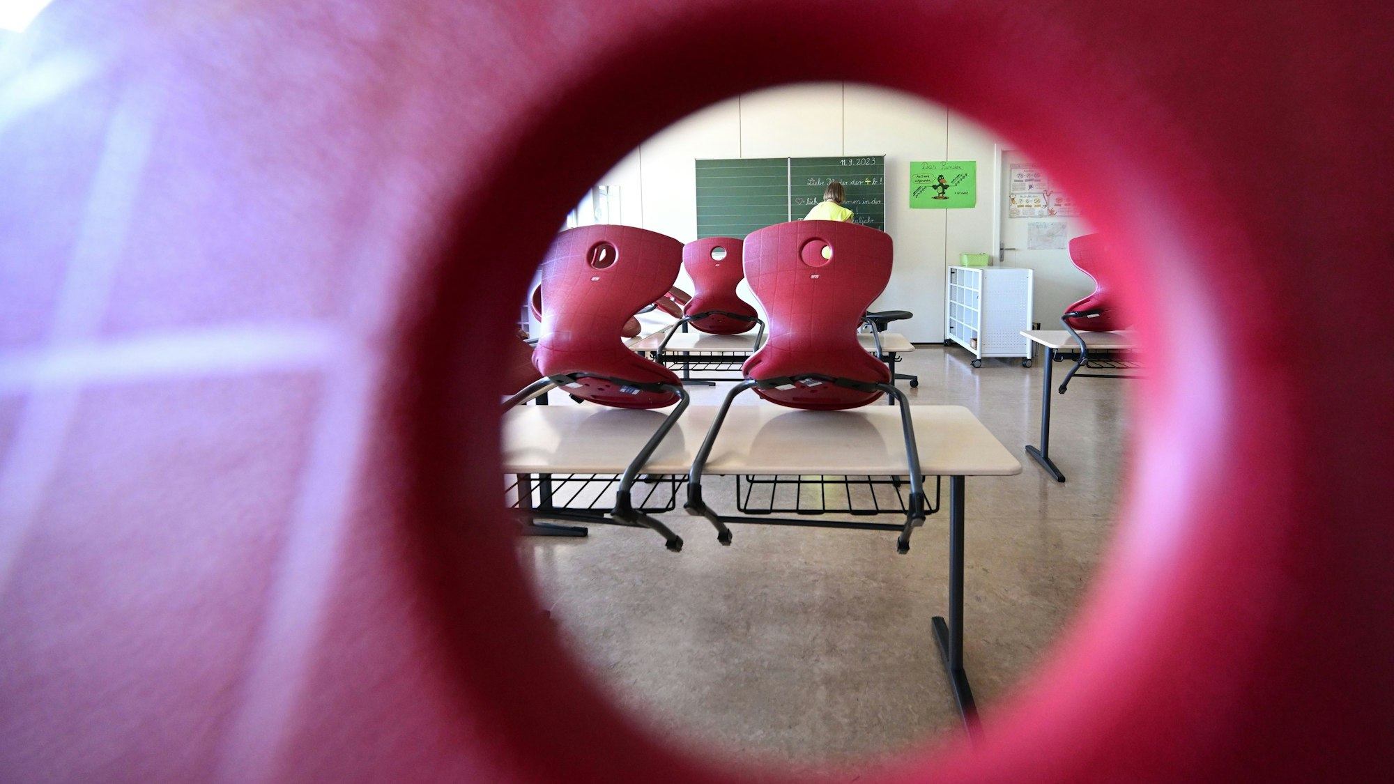 Stühle sind in einem Klassenzimmer auf Tische aufgereiht, im Hintergrund bereitet eine Lehrerin einen Tafelanschrieb vor.