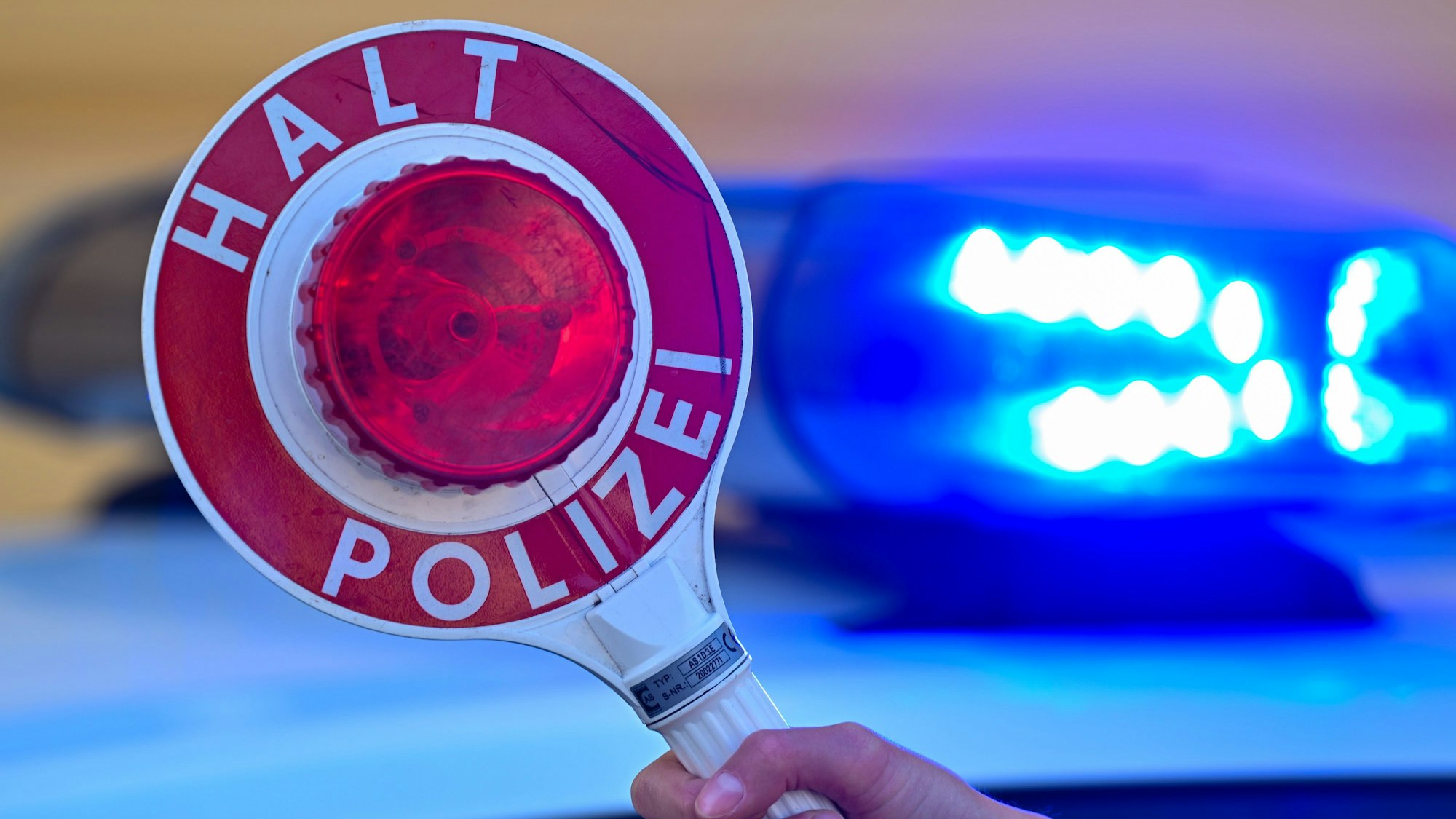 Eine Kelle der Polizei mit der Aufschrift Halt Polizei vor einem Blaulicht eines Polizeifahrzeuges.